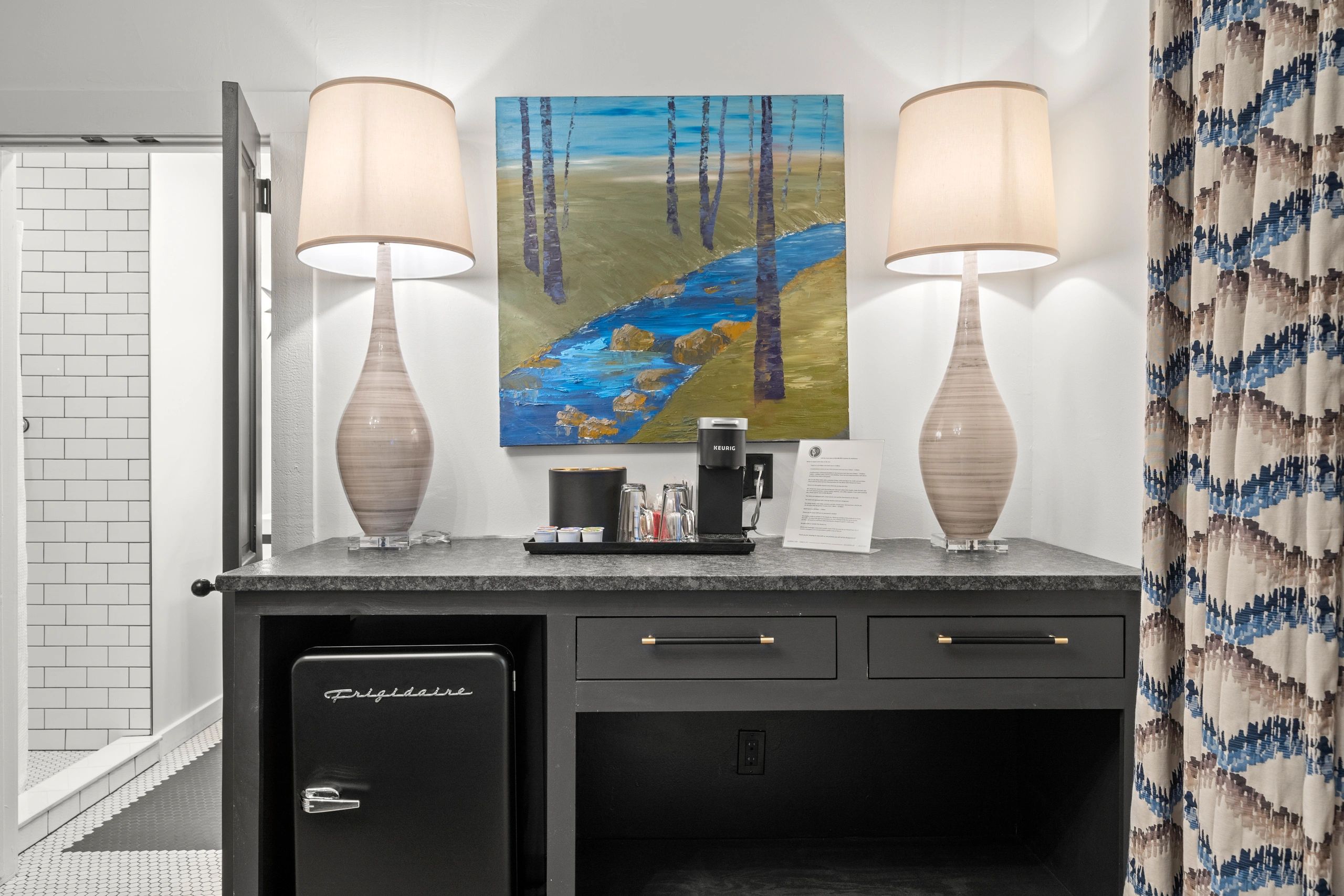 Hotel room desk area with two lamps, a painting, and a mini fridge